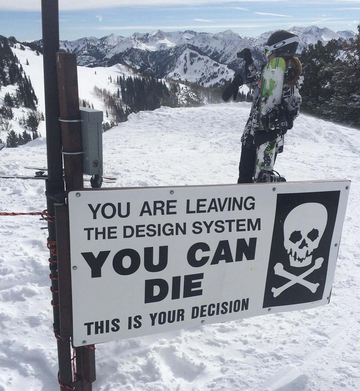 A photograph of a sign on a ski slope with a skull and bones which has been Photoshopped to say (in all caps) “You are leaving the design system. You can die. This is your decision.”
