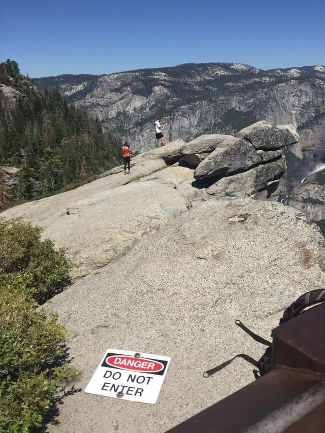 Photograph of a “DO NOT ENTER” sign on a rock cliff and people have passed it and are standing out on the edge of the cliff.