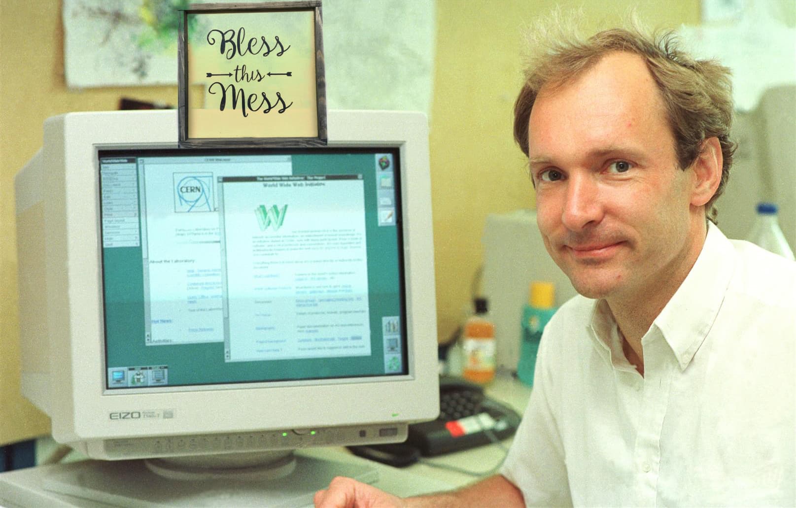 Photograph of Tim Berners-Lee next to his NeXT computer with the first web page on it at CERN. Above the image is a superimposed sign that says “Bless this mess” in cursive type.